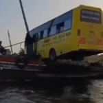 A school bus loaded onto a wooden boat floating on water in Lamu County, Kenya, surrounded by locals watching the transportation process.
